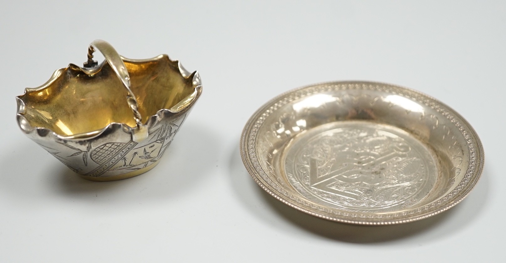 A late Victorian novelty parcel gilt silver salt modelled as a basket, with aesthetic engraved decoration, London, 1884, length 65mm, together with an Egyptian white metal small dish.